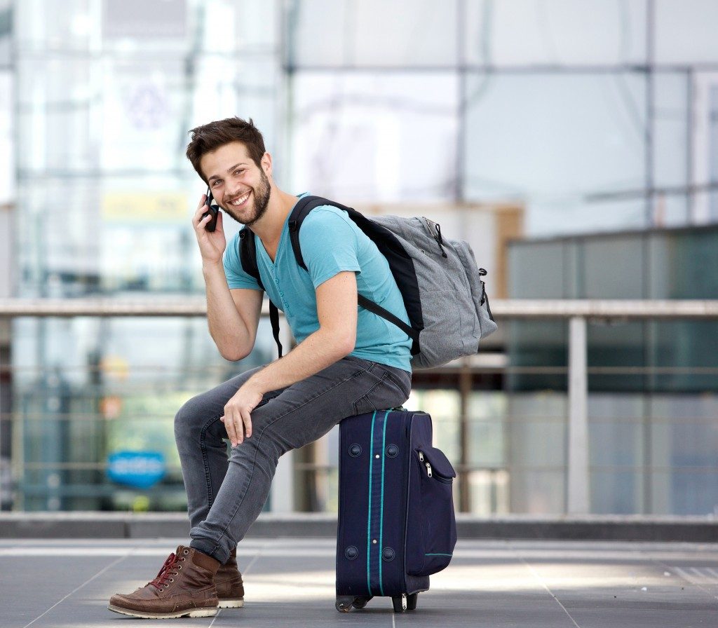 Portrait of a handsome young man sitting on suitcase and calling by cellphone at airport