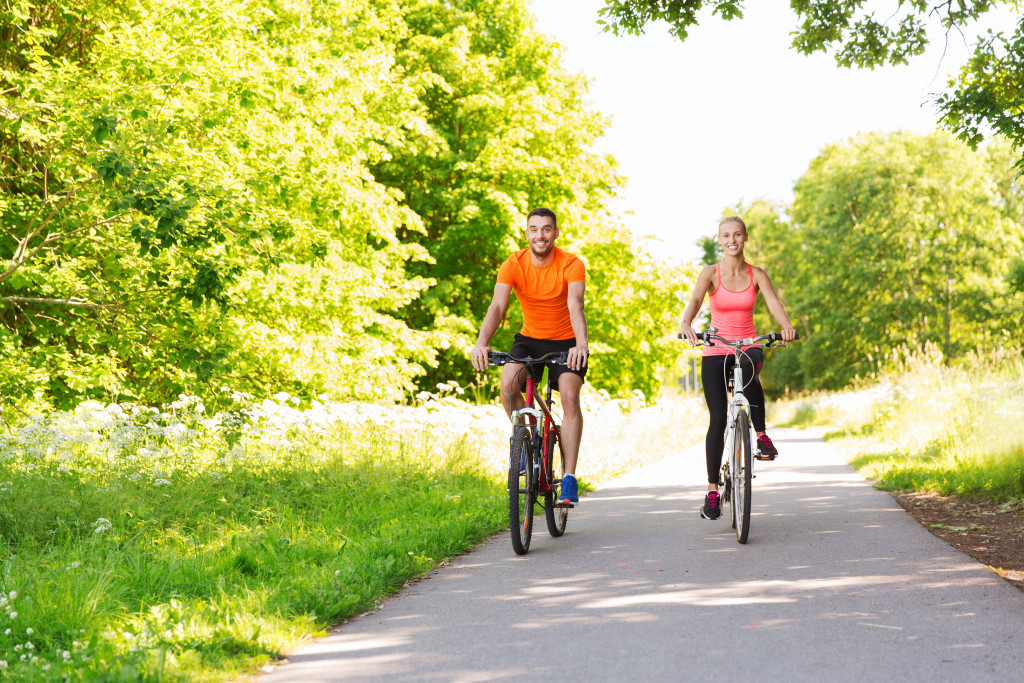 A male and female biking along a pavement surrounded by trees and grasses