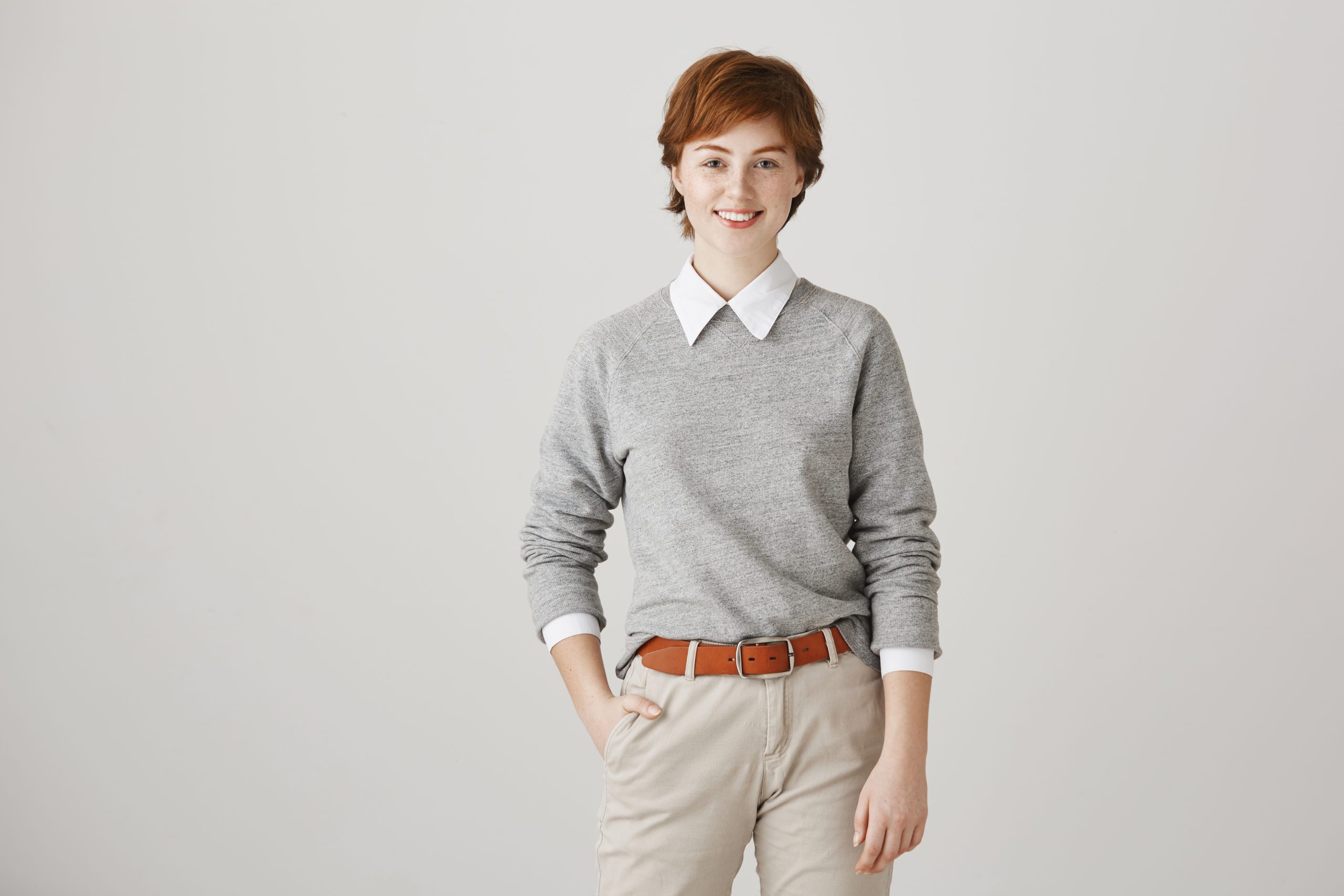 short-haired woman with one hand in her pocket, dressed in a gray sweater layered over a white collared shirt